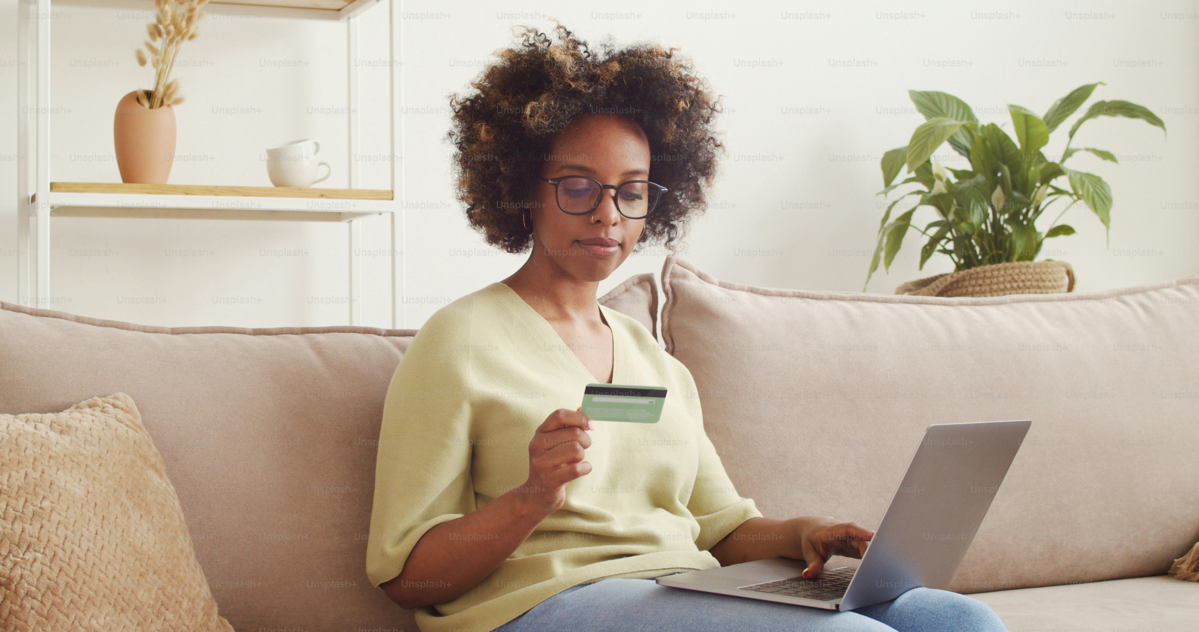 Young african woman paying online with credit card and laptop while sitting on sofa at home, buying clothes and shopping on ecommerce website